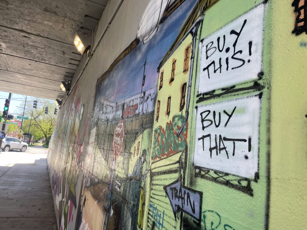 graffiti under a viaduct featuring a city scene, with an arrow point towards a train and signs that read BUY THIS! BUY THAT!
