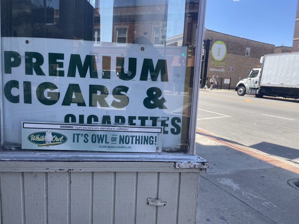a sign in a corner store window advertising premium cigars & cigarettes, with another ad for White Owl cigars reading "it's owl or nothing"