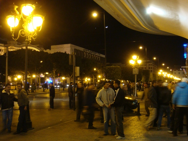 a crowded city street at night with rows of street lights and plenty of people talking.