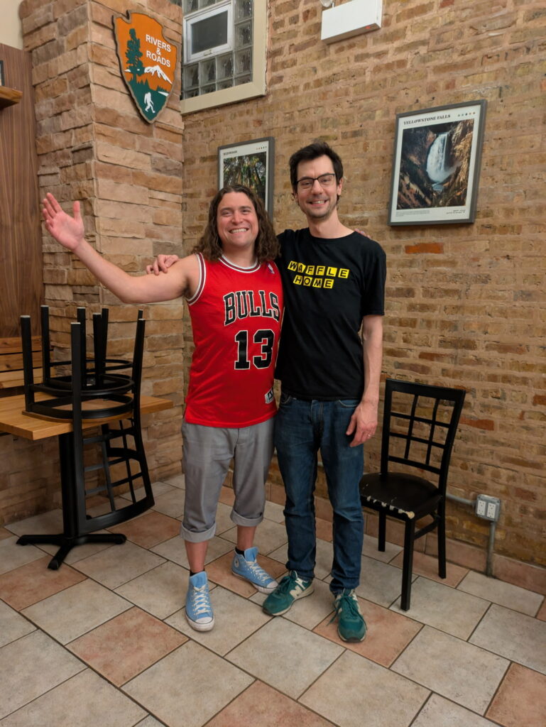 two men with their arms around each other smiling in a coffee shop with the chairs on the tables.
