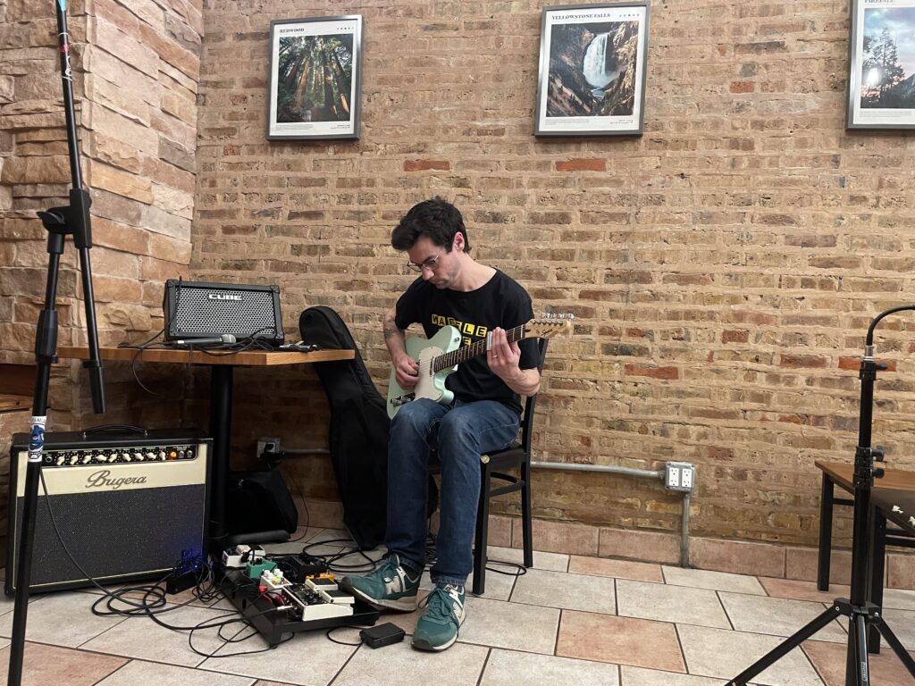 a man sitting in a coffee shop plays a blue Telecaster, eyes down on his pedalboard.