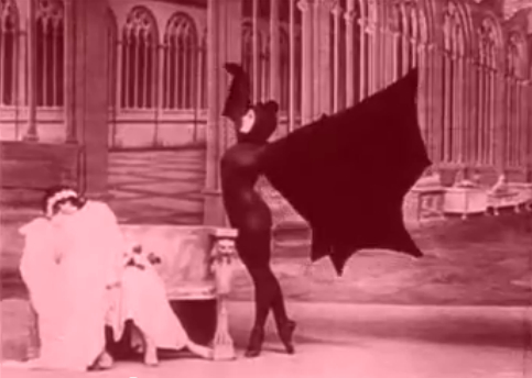 screenshot from a 1915 movie about vampires in which a woman in a black bodysuit and a black head covering and black bat wings towers over a cowering woman in white