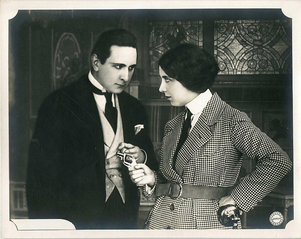 screentshot from an old black-and-white movie where a woman in a houndstooth trench coat holds a small gun to the chest of a man wearing a three-piece suit