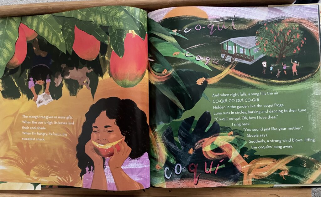 a shot of two pages of a children's book, depicting a young girl biting into a mango while underneath a mango tree on one page, and a house surrounded by swirling frogs on the other page