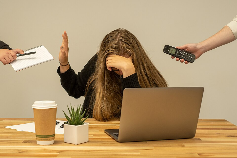 a woman sitting in front of a laptop, to-go coffee, succulent, and papers puts her head in her hands as people out of frame try to hand her a phone and a notebook with a pen.