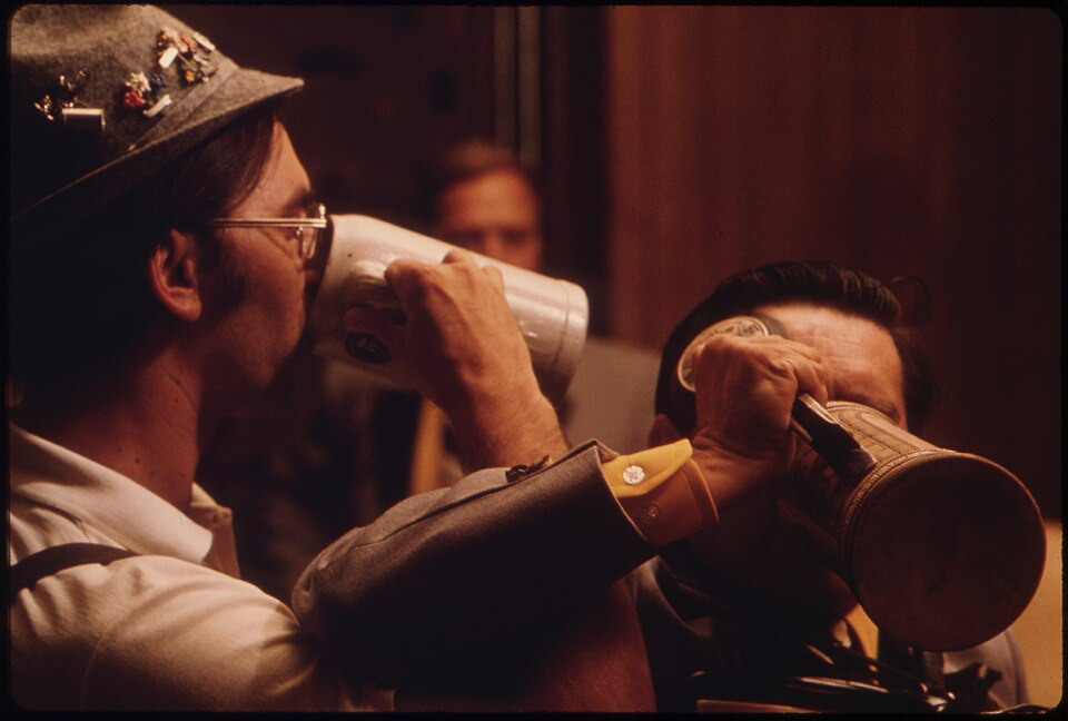 two men dressed in what appears to be Swiss yodeler Alpine gear drink deeply from oversized beer steins.