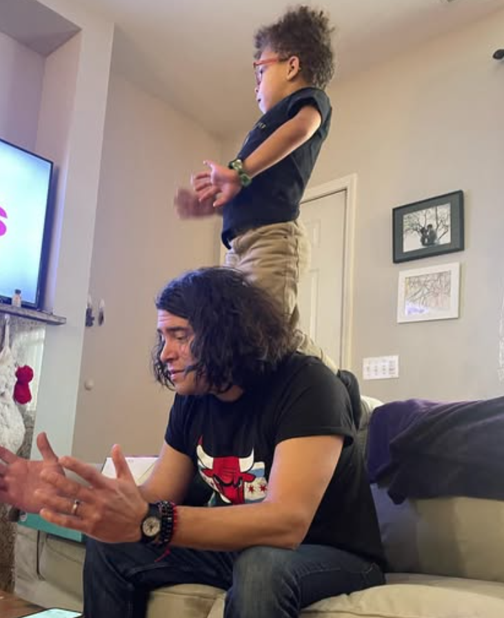 me on the couch, sitting upright and trying to talk, wearing yet another Chicago flag shirt with a Chicago Bulls logo over top of it. My then-five-year-old is sitting on his knees on my upper back, watching TV