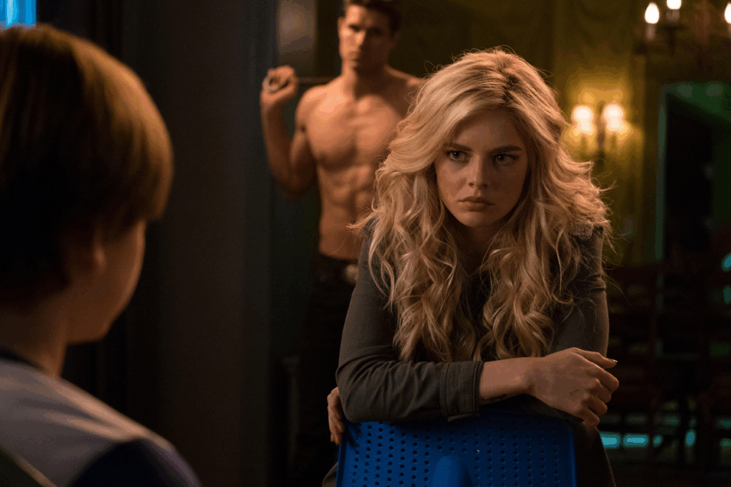 a blonde woman sits backwards in a chair, staring at a young boy, while a muscular man stands behind her with a baseball bat slung over his shoulder