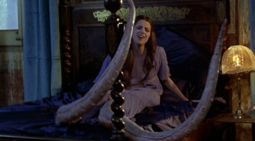 a woman sits on a bed, clothed and with a face twisted with either agony or desire or both, and both her legs are long tentacles