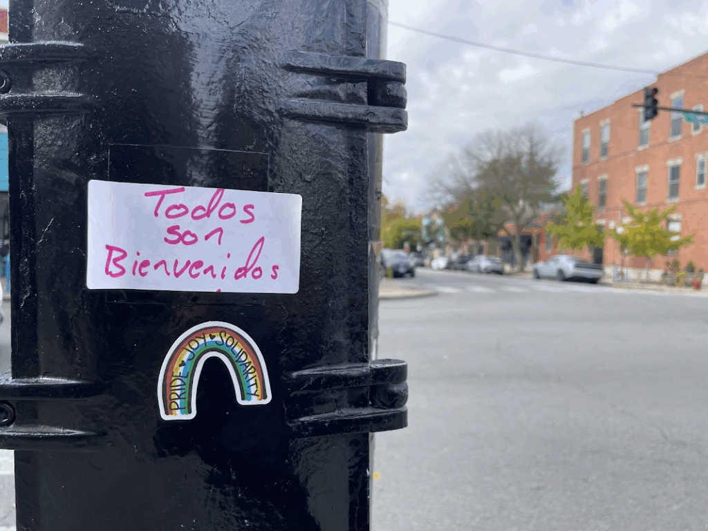 stickers on a black telephone pole, a white one with pink lettering reading todos son bienvenidos and a rainbow reading pride joy solidarity