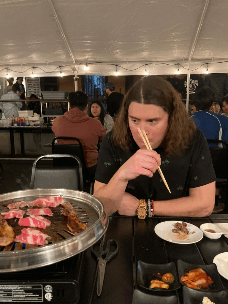 a white man with long brown hair and palm trees on his black shirt holds chopsticks and eyes pork belly and beef cooking on a Korean barbecue