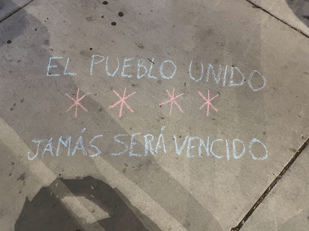 a chalk drawing on the sidewalk, in Chicago blue with red Chicago-style stars, reading EL PUEBLO UNIDO JAMÁS SERÁ VENCIDO