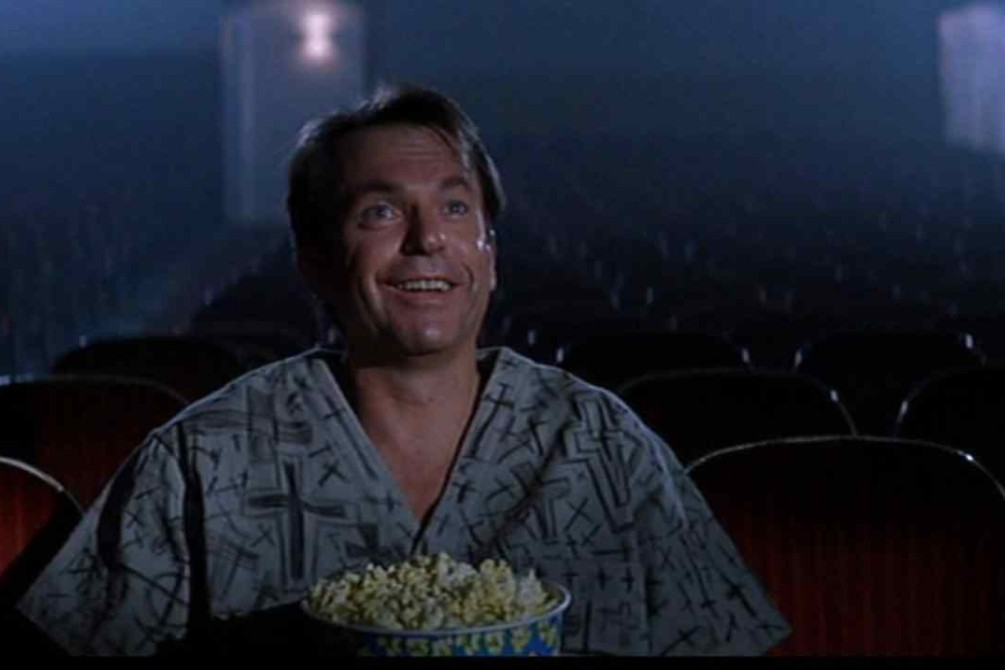 Sam Neil in the movie In The Mouth Of Madness, fully within said mouth, wearing the scrubs of an institutionalized person in a movie theater, smiling goofily at a movie with popcorn in his lap