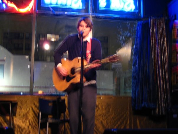 a man with mid-length brown hair wearing a black sweater and white shirt plays acoustic guitar in a shitty bar