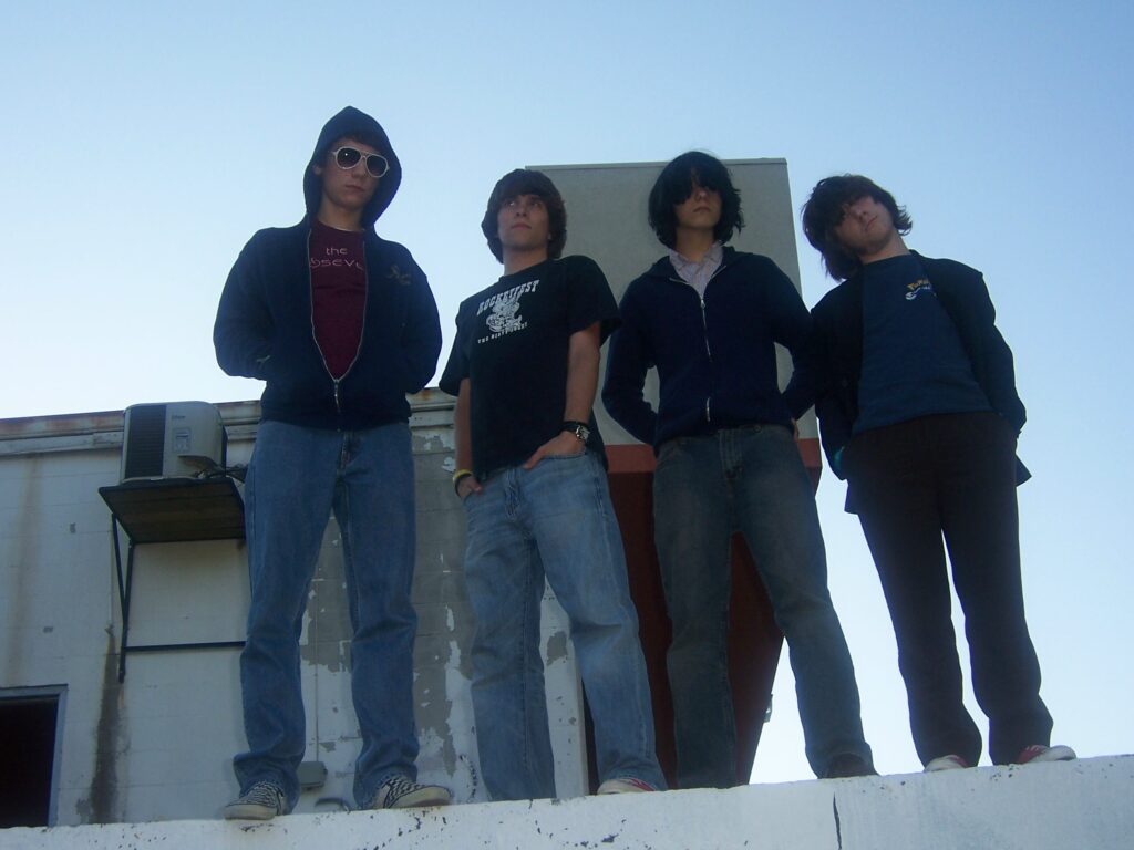 four white teenagers looking like if Frodo, Sam, Merry, and Pippin got really into the 2005 post-punk/emo scene, standing on a concrete wall