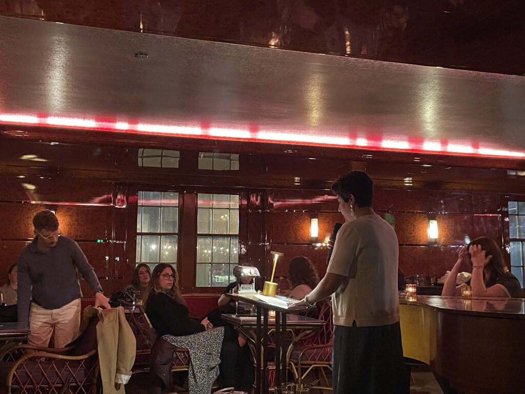 a brunette woman stands at a podium next to a piano in a dimly lit hotel bar, reading poetry into a microphone