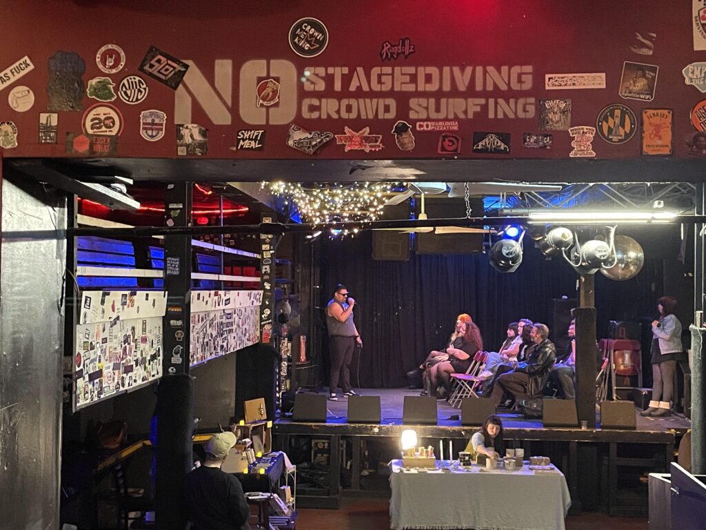 view of a stage with a person reading poems into a microphone, people sitting on stage, and a book and craft fair on the level below the stage. In the foreground, the band sticker-covered wall has a stenciled NO STAGEDIVING CROWD SURFING