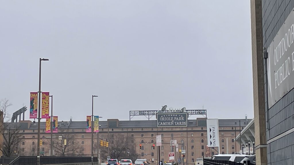 a street view of a brick baseball stadium with a black sign with orange lettering reading Oriole Park at Camden Yards