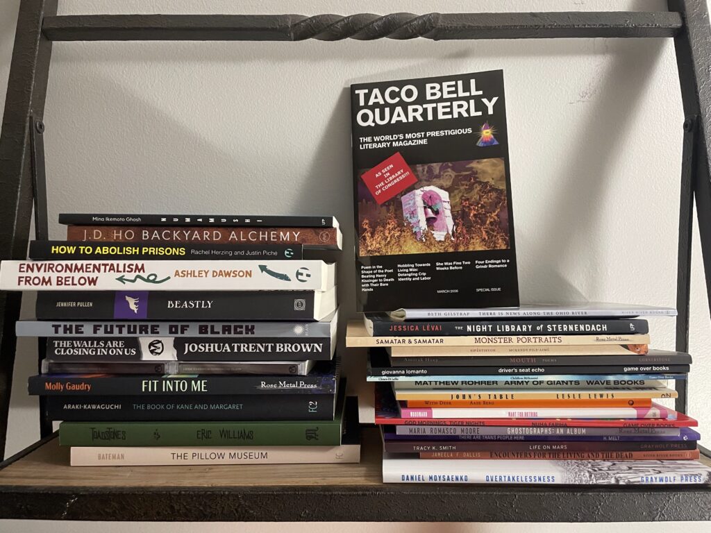 a pile of paperback books on a shelf, with the print issue of Taco Bell Quarterly prominently displayed