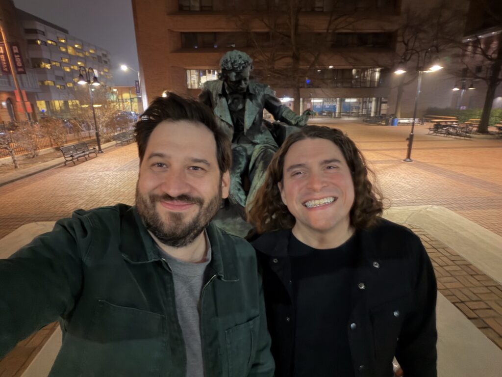 two white men in jackets, one bearded and one a longhair, smiling in front of a statue of Edgar Allen Poe in downtown Baltimore