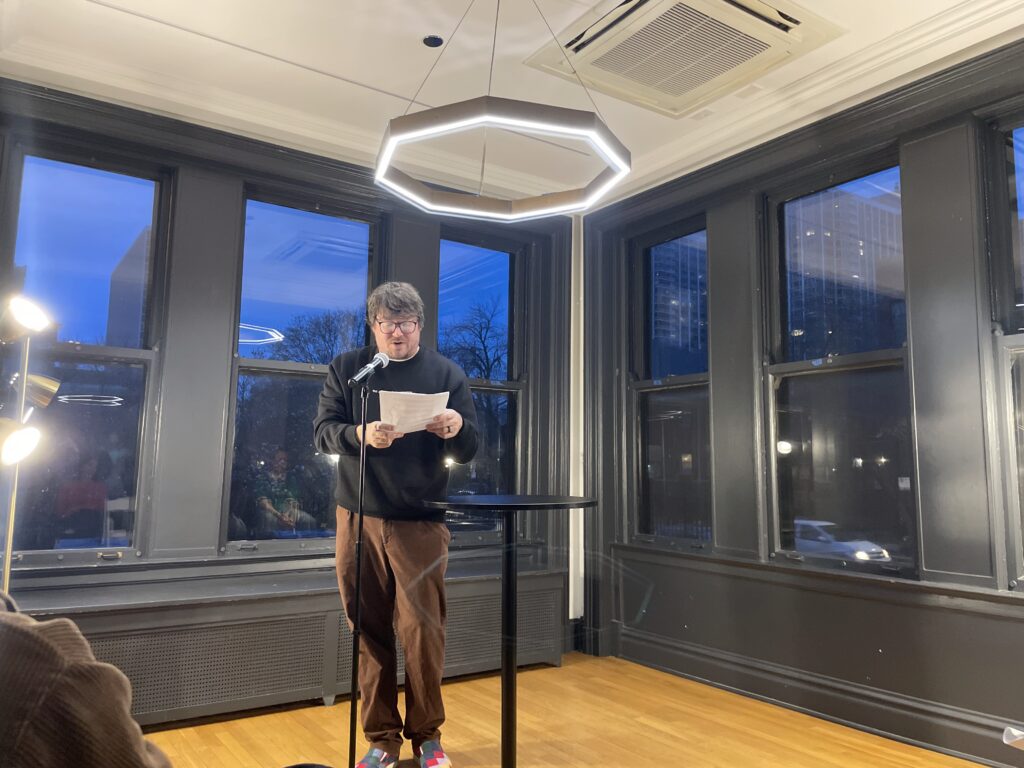 a man in a sweater reading at a microphone