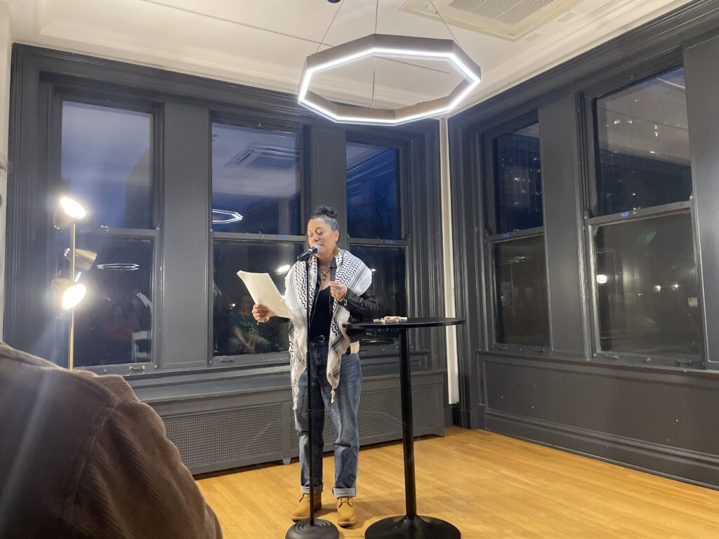 a woman wrapped in an incredible patterned scarf, black shirt and jeans, and some impressive rings reads at a microphone
