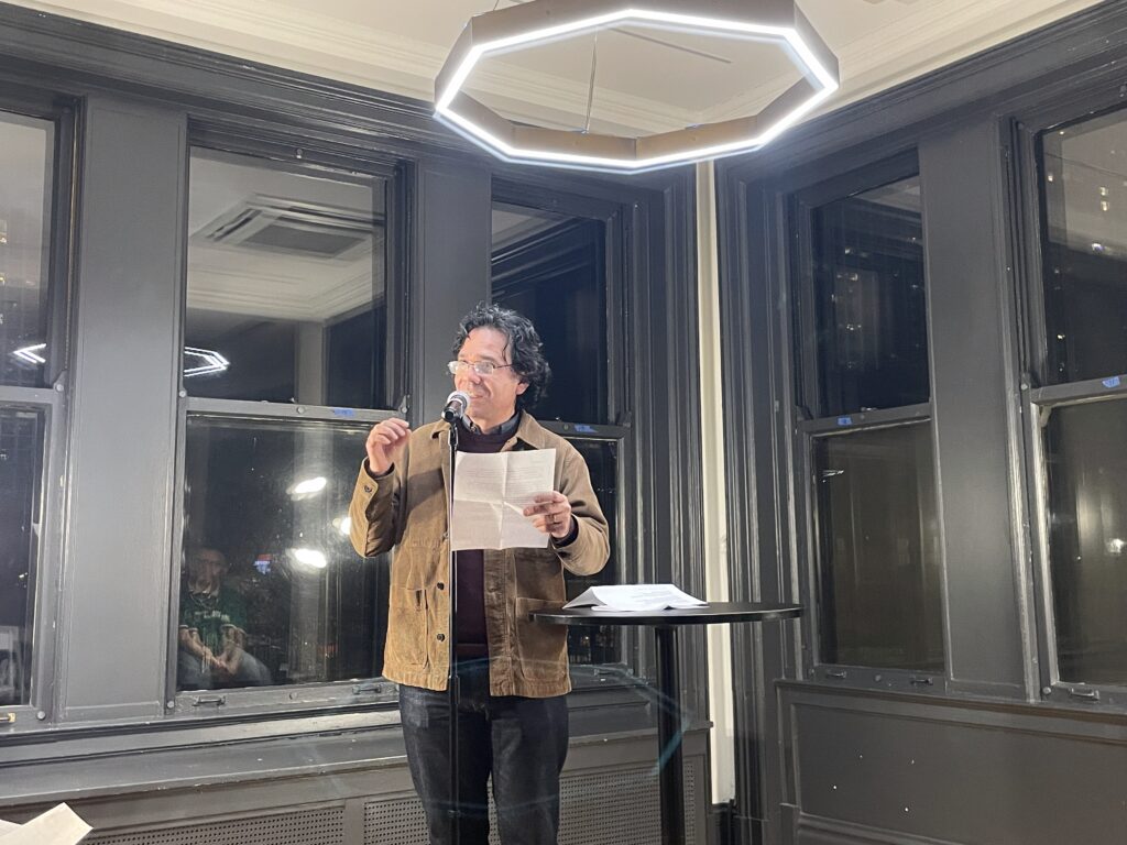 a man with incredible, thick hair and a cool professor's jacket reads at a microphone