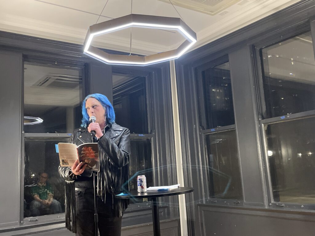 a poet dripping in swag with a fringed leather jacket, black jeans, a spiked bracelet, and shoulder-length blue hair reads at a microphone