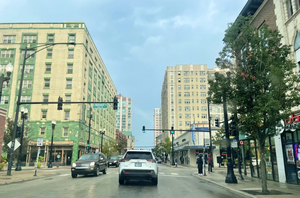 photo of Bryn Mawr ave looking east from the train station. Winthrop is visible, it is drizzling a bit, the green affordable housing building and Hamburger Mary's (née That Little Mexican Cafe) are visible, people are on the sidewalks.