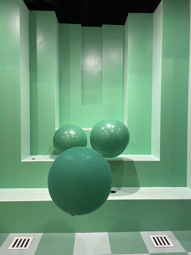 an art installation with ascending green stairs to kind of look like a collapsing square, plus three big balloons floating