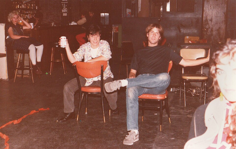 two people with brown hair and decent but unremarkable clothes sit on red chairs at a punk club, one person toasts a Budweiser